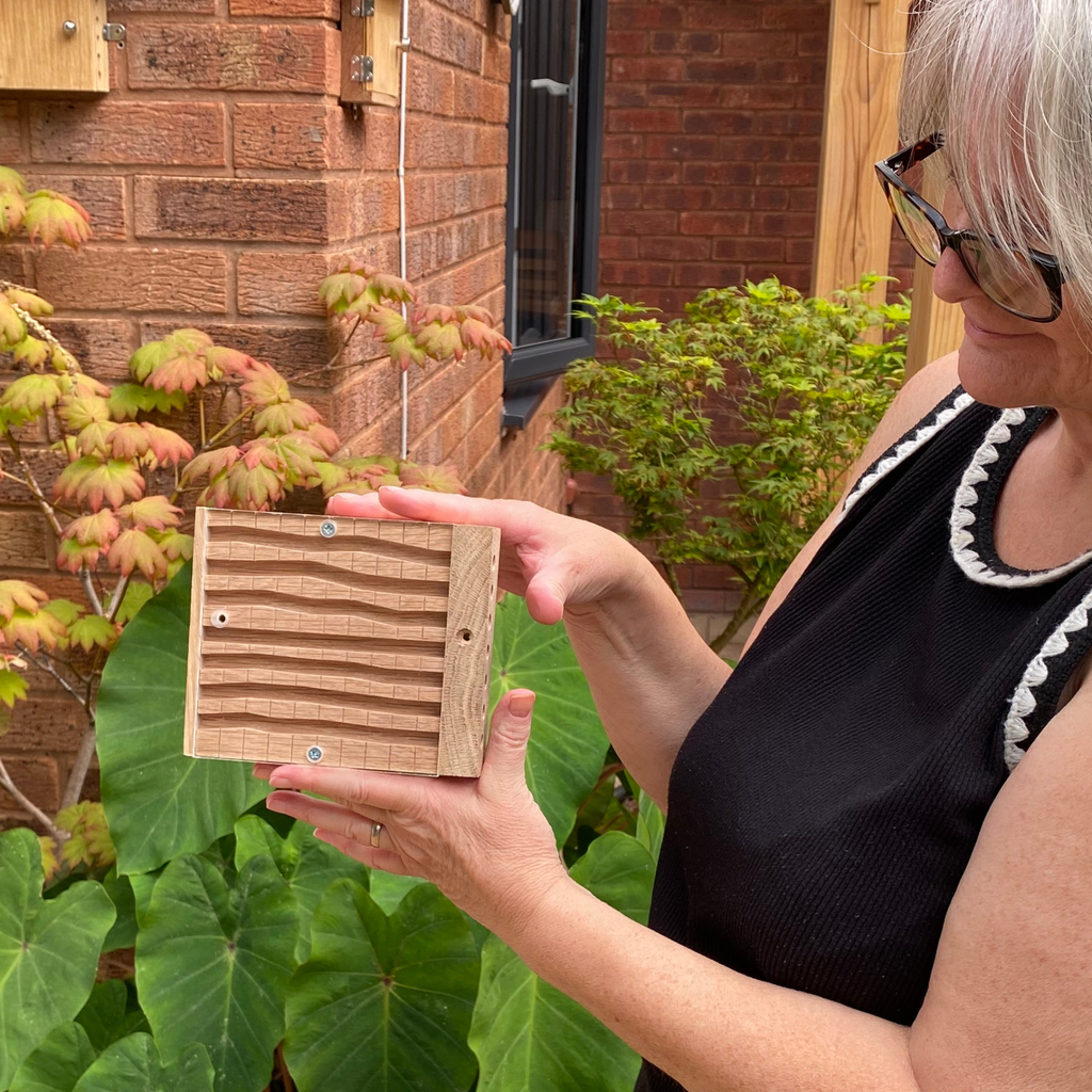A woman outdoors in a garden setting holds a small wooden box with a slatted front and visible screws. The background shows brick walls, green plants, and a window.